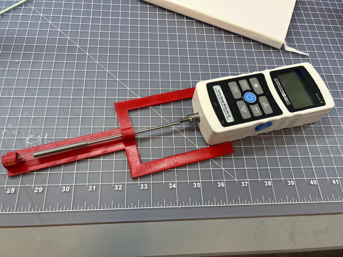 Red 3D-printed fixture holding a steel cannula, connected to a Mark-10 force gauge on a cutting mat