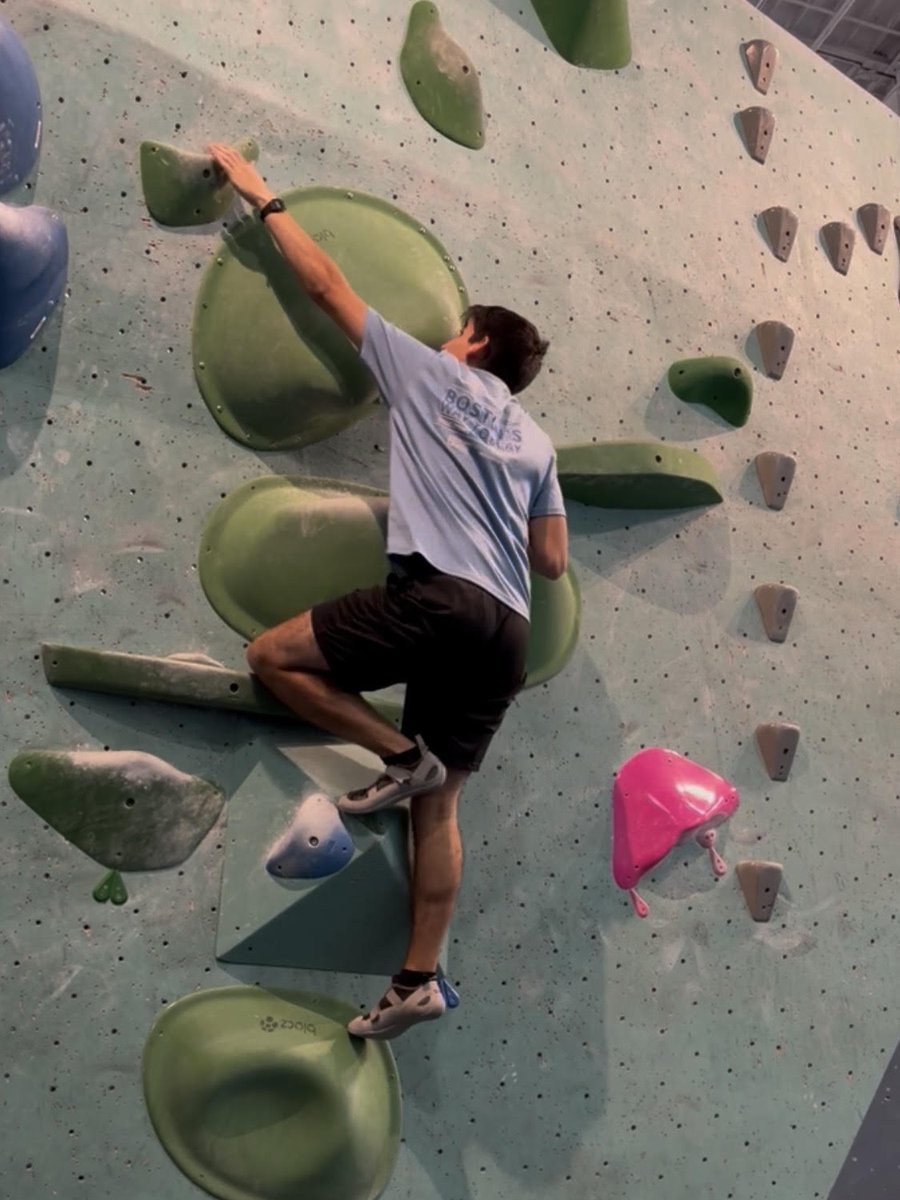 Jordon mid-climb on a bouldering wall, reaching for a green hold