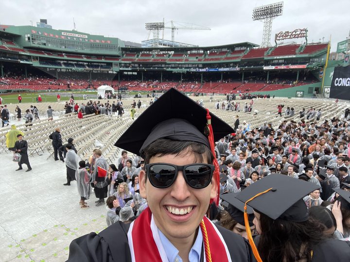 Jordon in cap, gown and sunglasses taking a selfie with fellow graduates at Fenway Park