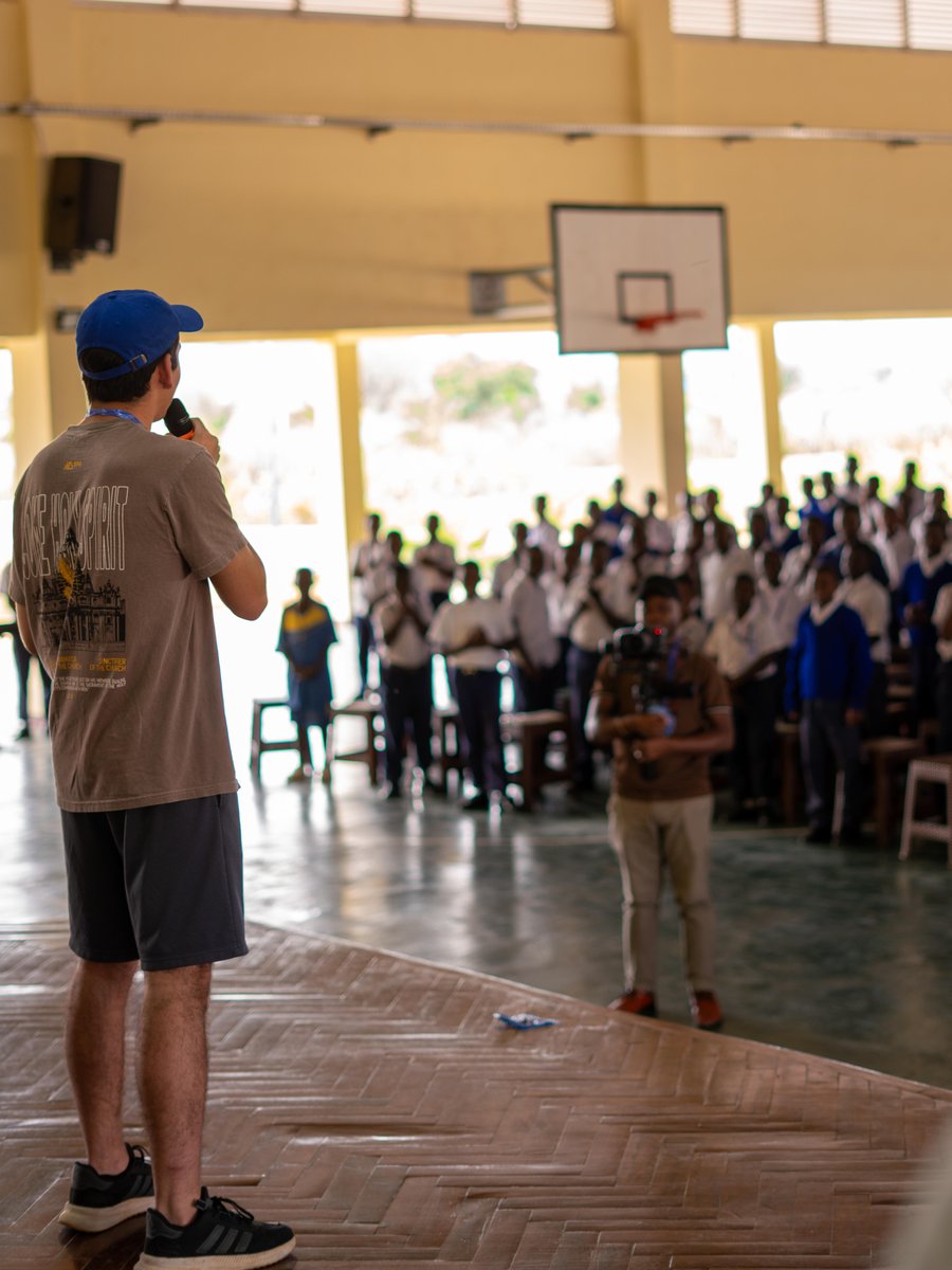 Jordon speaking into a microphone to a gym full of students in school uniforms in Tanzania